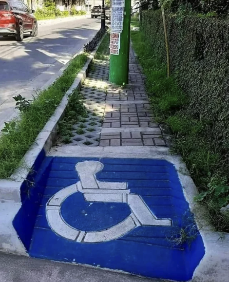 A handicapped blue/white sign on the ground at the end of a narrow sidewalk that has - not far ahead of it - a pole ( that a wheelchair couldn’t go around unless you go up and walked around it ) and uneven breaks.