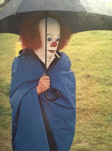 Clown from IT (Tim Curry) with a cigarette in it's mouth holding an umbrella in light rain with a blue blanket wrapped around him. Tim is just starring up into the distance.