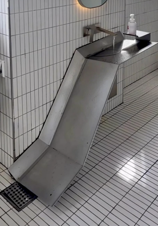 A metal sink in a bathroom with one side that extends down like a waterfall slide into a drain on the floor instead of having a pipe take the water away.