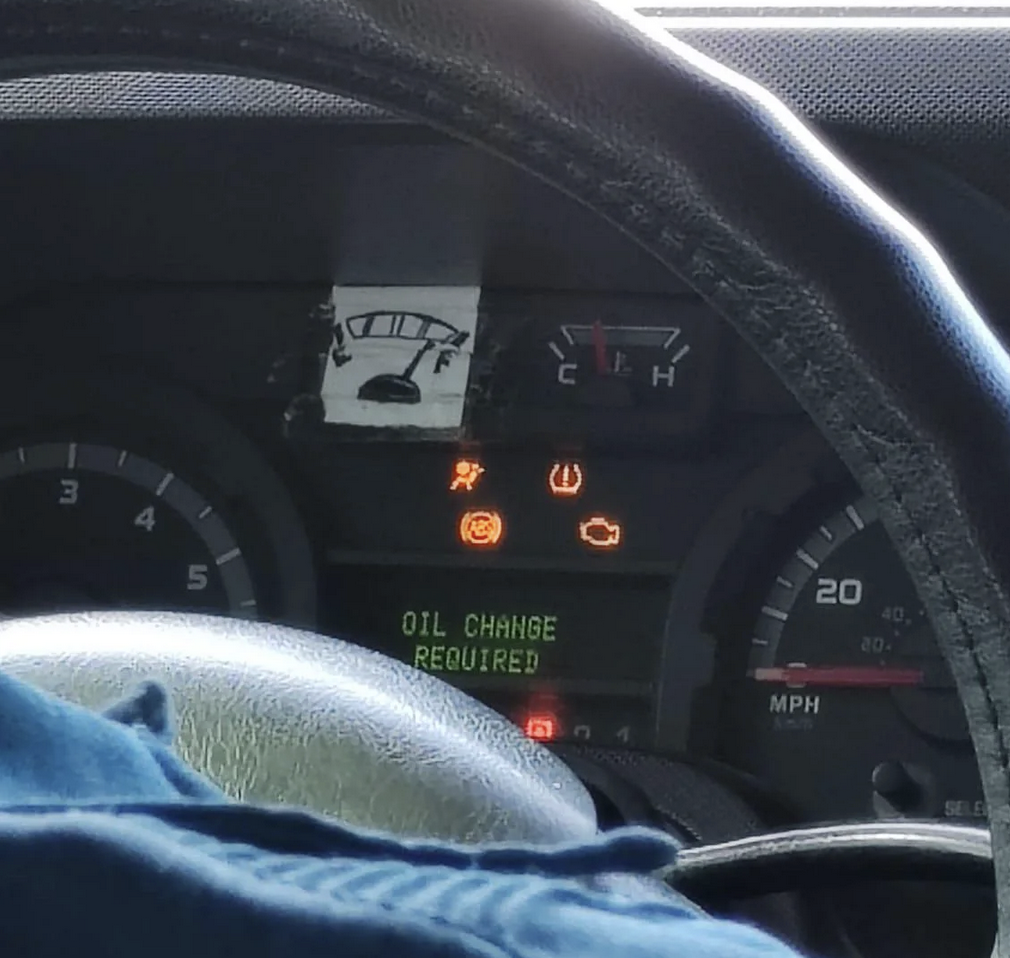 Dashboard of a driver who has many warning lights on and a "oil change required" on a digitial display on the dash as well. Person has drawn a full gas tank indiactor and pasted it over the real one.