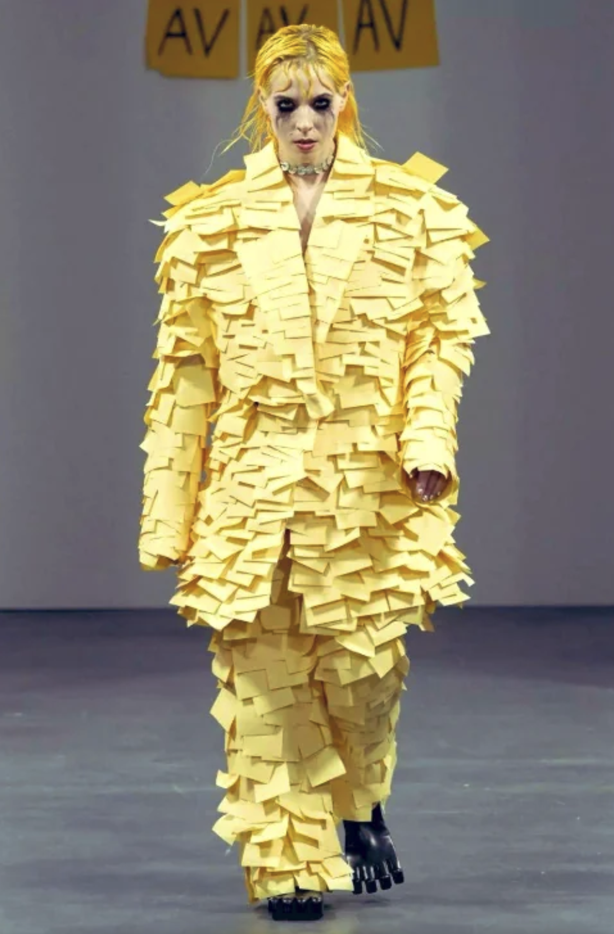 What seems to be a yellow haired woman walking down a hallway or runway in an outfit made head to toe with seemingly post it notes.