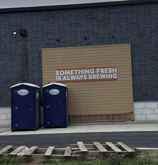 Outside on a wall the slogan from coffee shop: "Something Fresh is Always Brewing" with two porta potties off to the site of it.