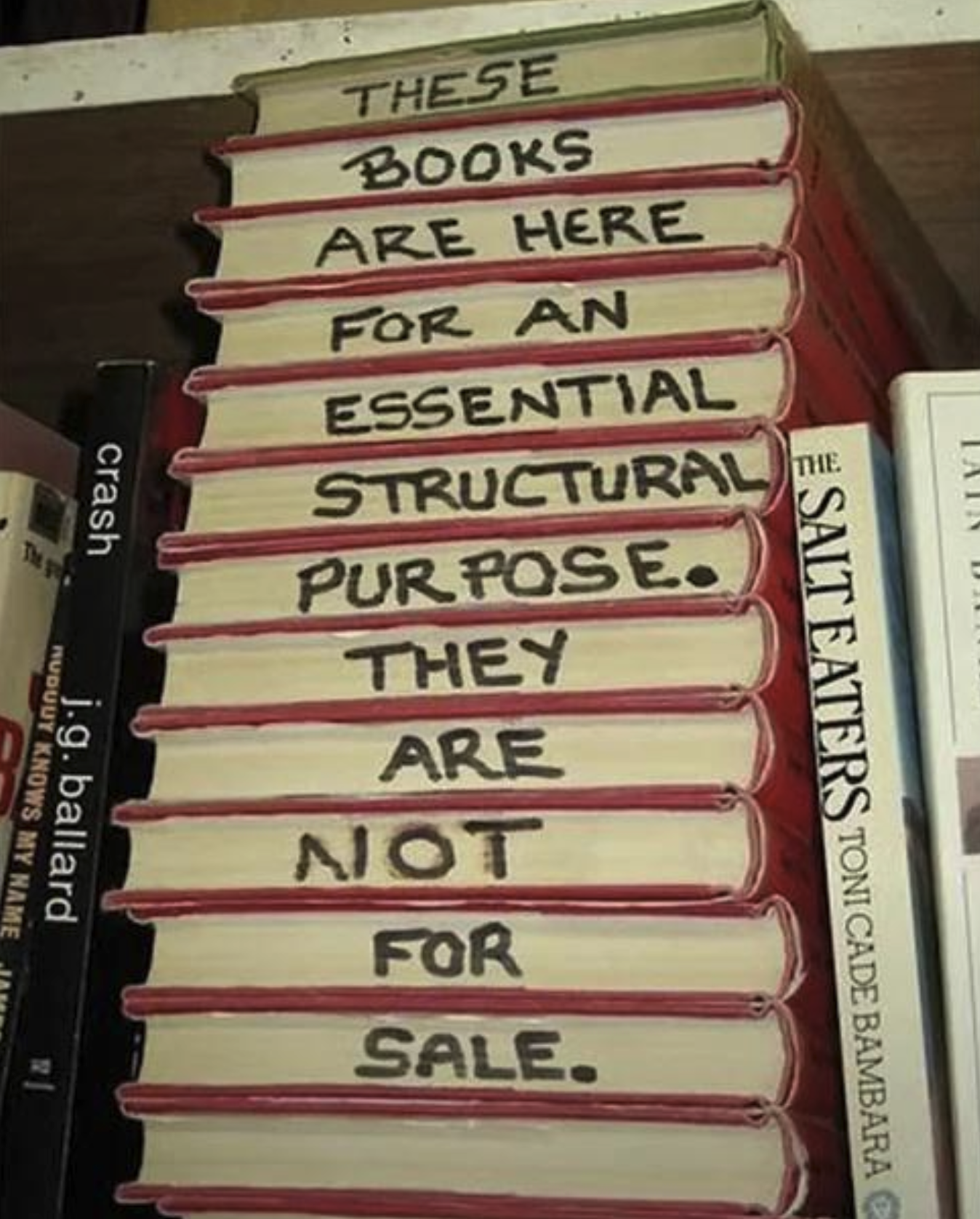 Stack of books on a bookshelf holding it up with the words "These books are here for an essential structural purposes. They are not for sale" written on the sides of the books from top to bottom.