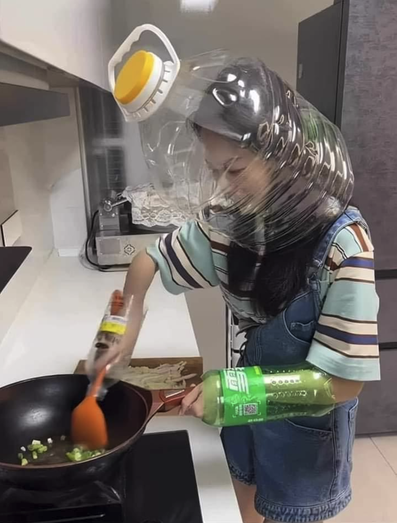 Woman cooking on a stove top but us using empty plastic water bottles for mittens and her had is in an empty transparent water jug to protect her face/head from the cooking oil in the pan.